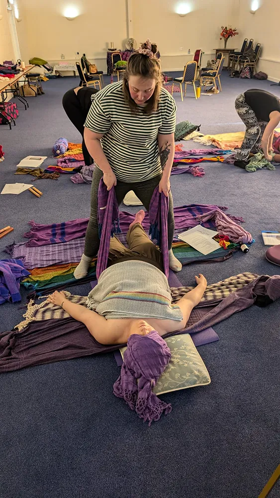 Doula support A rebozo workshop where Zoe gently rocks a person lying on the floor, wrapped in scarves