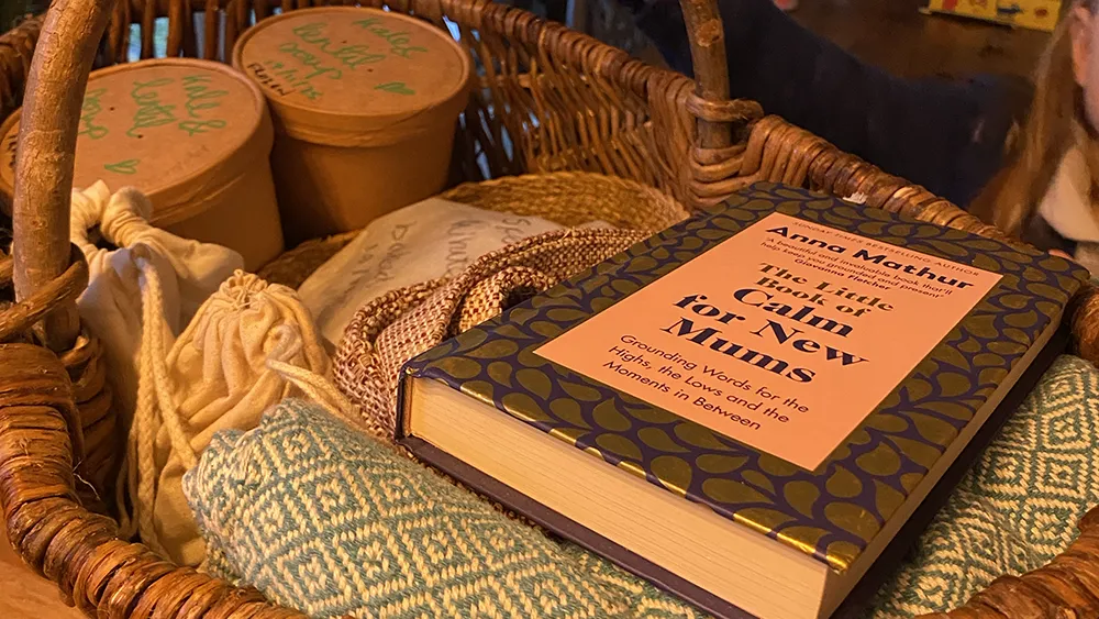 book-in-hamper A wicker hamper holding a copy of "Calm for New Mums", a woven blanket and tubs of food