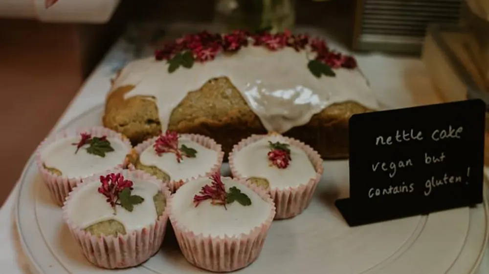 nettle-cake A nettle cake and cupcakes decorated with rose petals, with a sign saying it's vegan but contains gluten
