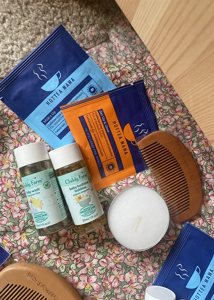 Postpartum care items: herbal bath teas, a wooden comb, baby wash and a candle on a floral blanket