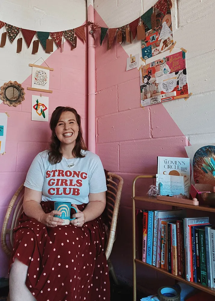 Zoe is sat holding a mug Zoe smiling, sitting in a wicker chair holding a mug in front of a pink wall and bookshelf